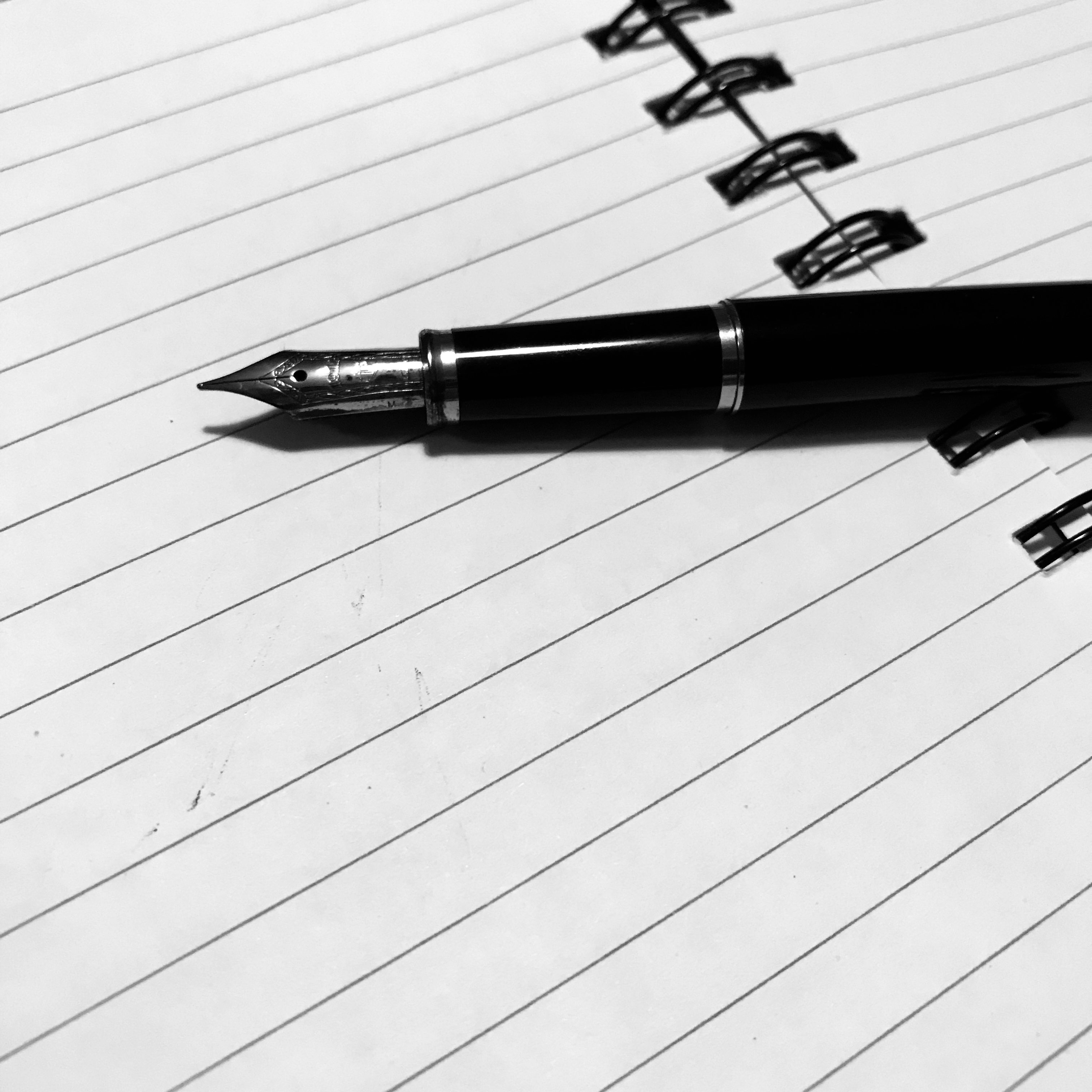 A black and white photo of a black photo pen with a shiny nib lying on top of a the middle of a lined, spiral-bound notebook - the lined pages of the notebook are blank 
