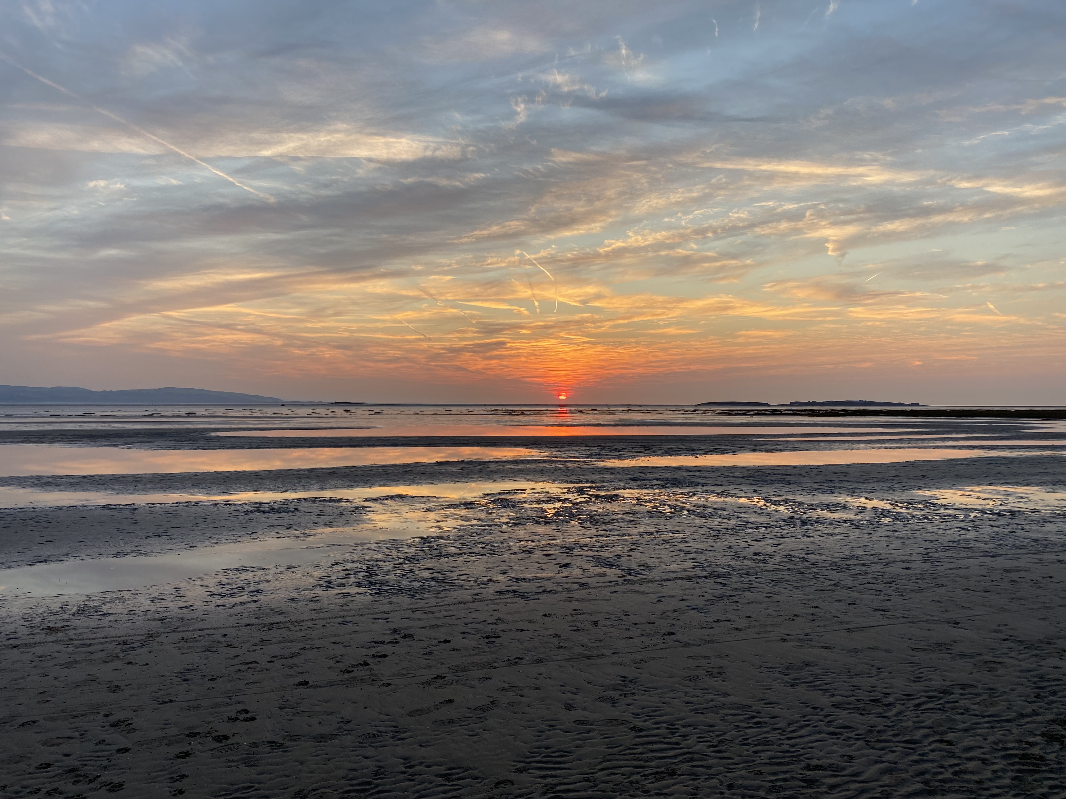 A red sun sets on the horizon of a beach, the red rays of the sun reflected in the clouds and back on the puddles of water on the beach. The north headland of Wales is visible in the distance